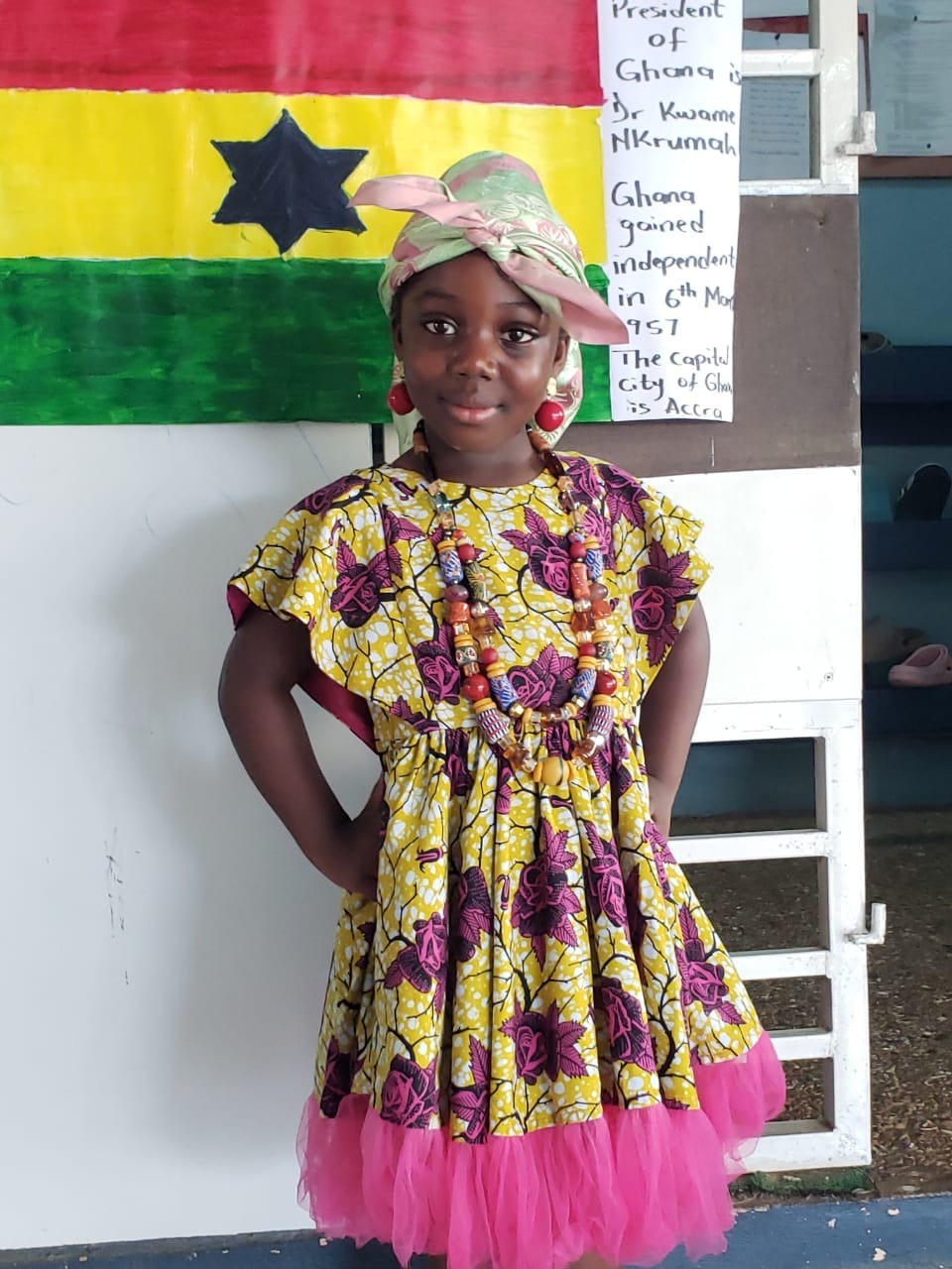 Girl in traditional Ghanaian kente dress and headwrap posing in front of the Ghana flag