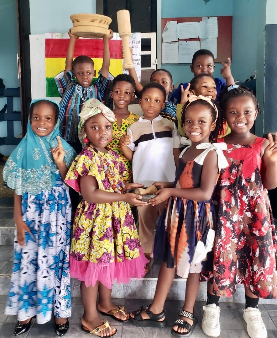 Students in traditional Ghanaian attire holding a mortar and pestle in front of the Ghana flag