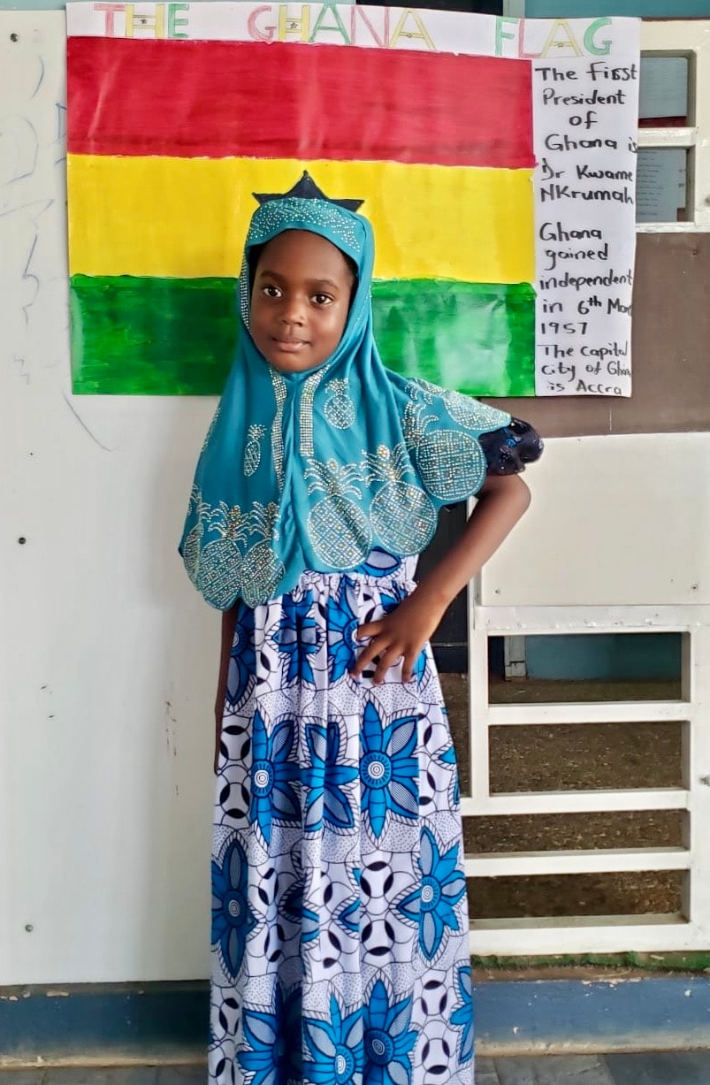 Girl in embroidered blue hijab and kente skirt posing in front of the Ghana flag